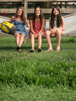 Tres adolescentes sonrientes disfrutando de su primer campamento de verano, sentadas en una hamaca, representando la experiencia y los amigos en los campamentos.