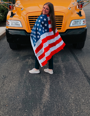 Estudiante sonriente frente a un autobús escolar con una bandera, representando la experiencia de transformación de un año académico en el extranjero.